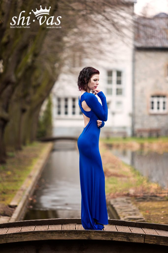 Coco steht im Profil auf einer Holzbrücke über einem Kanal; das blaue Kleid betont ihre gesamte Rückansicht und die schmale Silhouette.