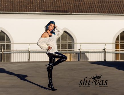 Coco steht frontal auf einem Parkhausdach, trägt eine glänzend schwarze Latex-Leggings und einen weiten Pullover, ein Bein leicht angewinkelt, fotografiert im warmen Herbstlicht.