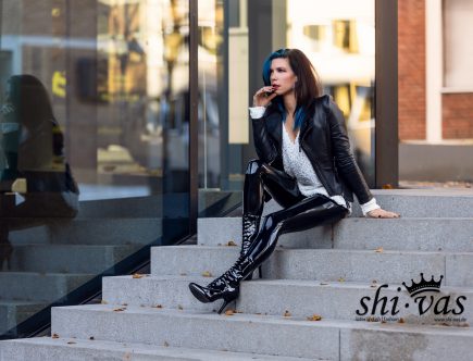 Coco sitzt auf den Stufen vor einer spiegelnden Glasfront, Hand am Kinn, schaut nachdenklich zur Seite, ihre Spiegelung ist im Fenster zu sehen