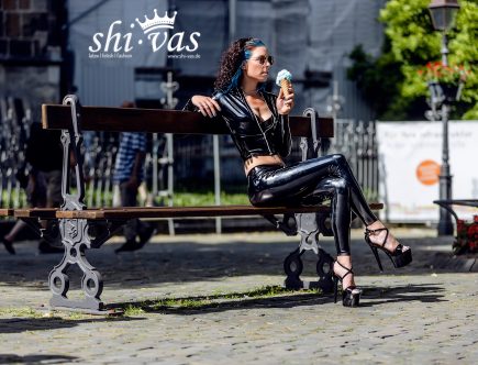 Coco sitzt auf einer Parkbank, Beine elegant übereinandergeschlagen, hält ein Eis in der Hand, schaut zur Seite, Ganzkörperaufnahme.