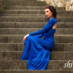 Coco sitzt elegant auf einer breiten Steintreppe; das bodenlange blaue Kleid umspielt ihre Beine, während sie nachdenklich zur Seite blickt.