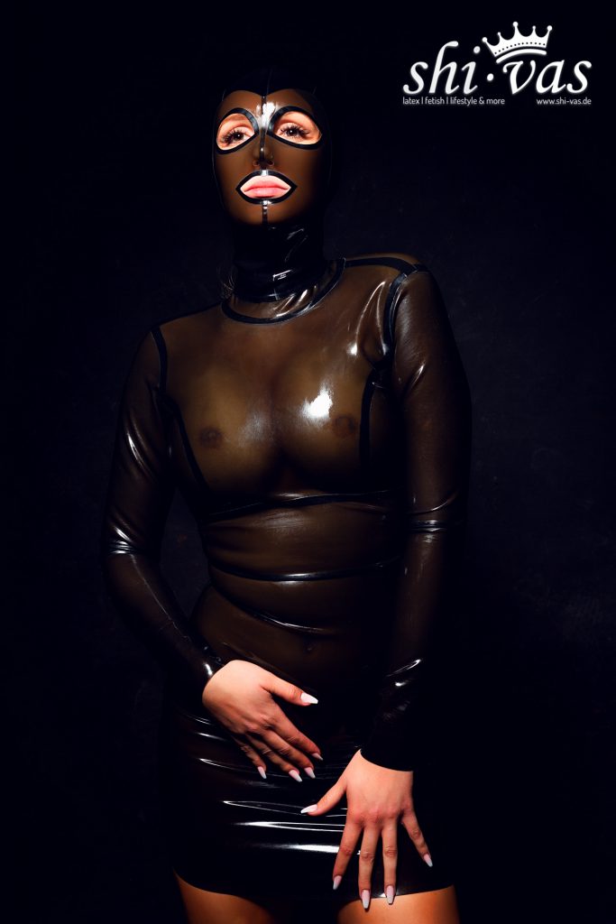 Finnja steht frontal in einem semi-transparenten, braunen Latexkleid mit schwarzer Maske. Ihre Hände liegen auf ihren Oberschenkeln.