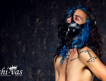 Extreme Nahaufnahme. Kopf im Nacken, Augen geschlossen, der Penisknebel sitzt fest. Der Ausdruck schwankt zwischen Schmerz und höchster Lust.