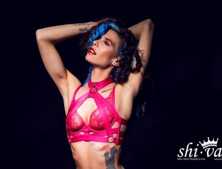 Coco steht frontal vor schwarzem Hintergrund, beide Arme hinter dem Kopf verschränkt, der Oberkörper offen.