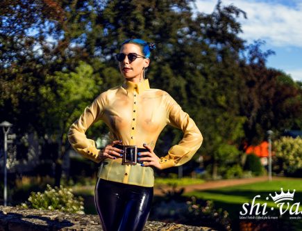Coco steht vor einer grünen Hecke, trägt eine naturtransparente Latexbluse, eine schwarze Latex-Leggings und Sonnenbrille. Die Hände hat sie in die Taille gestützt.