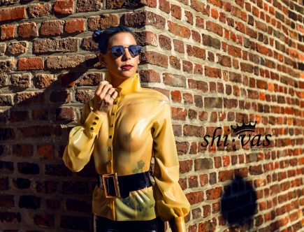 Coco steht eng an der Mauer, eine Hand am Kragen der Bluse, blickt direkt und intensiv in die Kamera.