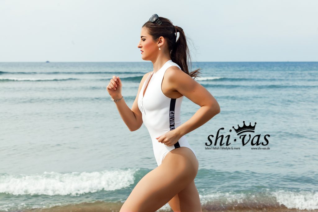 Lina läuft im weißen Calvin-Klein-Onepiece am Strand entlang. Der nasse Stoff betont ihre Körperlinien, Bewegung und Präsenz verbinden sich zu klarer, selbstbewusster Erotik.