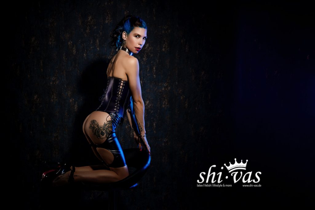 Fetisch-Model Coco in schwarzem Latex-Korsett und Strümpfen, kniend mit intensivem Blick in die Kamera, sinnlich und selbstbewusst inszeniert vor dunklem Hintergrund