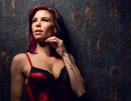 Coco steht vor einem dunklen, strukturierten Studiohintergrund und trägt rote Dessous. Ihr Blick ist leicht nach oben gerichtet, die Pose aufrecht und selbstbewusst, das Licht betont Gesicht und Oberkörper.