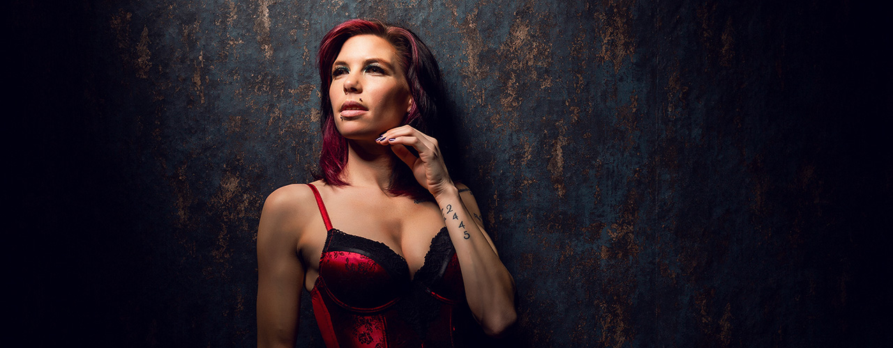 Coco steht vor einem dunklen, strukturierten Studiohintergrund und trägt rote Dessous. Ihr Blick ist leicht nach oben gerichtet, die Pose aufrecht und selbstbewusst, das Licht betont Gesicht und Oberkörper.