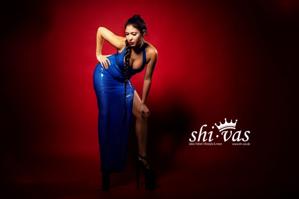 Gastmodel Salome beugt sich in einem semitransparenten blauen Latex-Abendkleid mit hohem Beinschlitz und extrem hohen Heels nach vorne, roter Studiohintergrund.