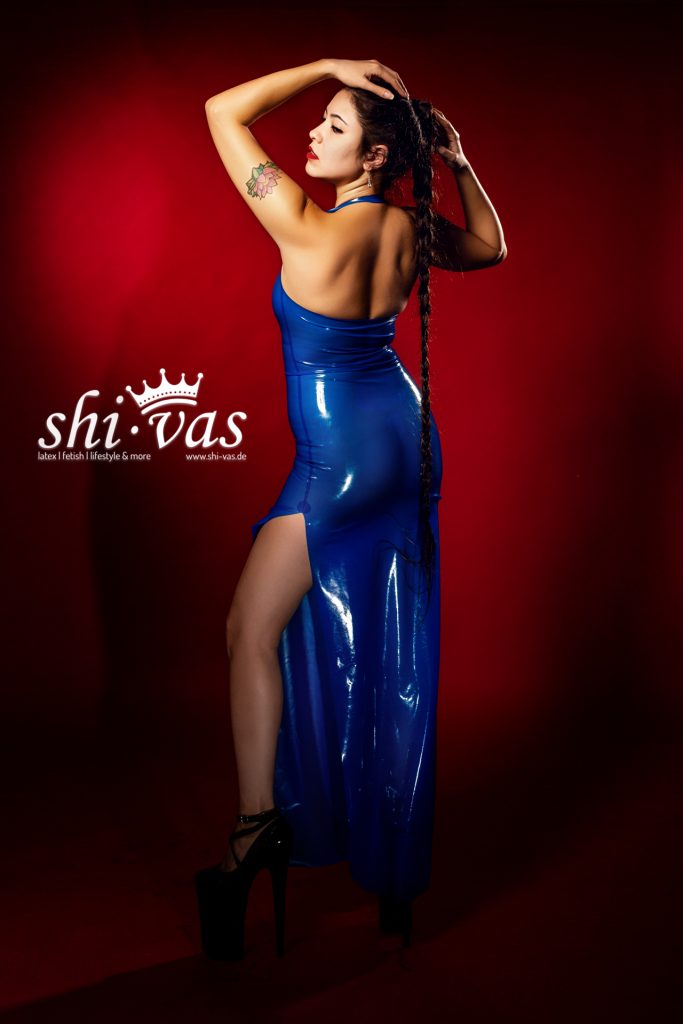 Gastmodel Salome von hinten in einem semitransparenten blauen Latex-Abendkleid mit hohem Beinschlitz und extrem hohen Heels vor rotem Studiohintergrund.
