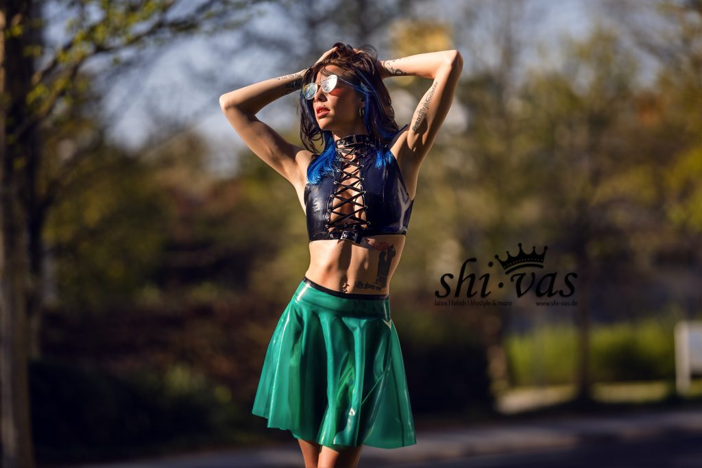 Coco steht im Sonnenlicht mit erhobenen Armen, trägt ein schwarzes geschnürtes Latex-Top und einen semi-transparenten grünen Latexrock ohne Slip. Das Latex glänzt im Licht, entspannte aber selbstbewusste Pose, urbaner Hintergrund, sinnliche Spannung.