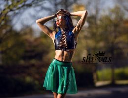 Coco steht im Sonnenlicht mit erhobenen Armen, trägt ein schwarzes geschnürtes Latex-Top und einen semi-transparenten grünen Latexrock ohne Slip. Das Latex glänzt im Licht, entspannte aber selbstbewusste Pose, urbaner Hintergrund, sinnliche Spannung.