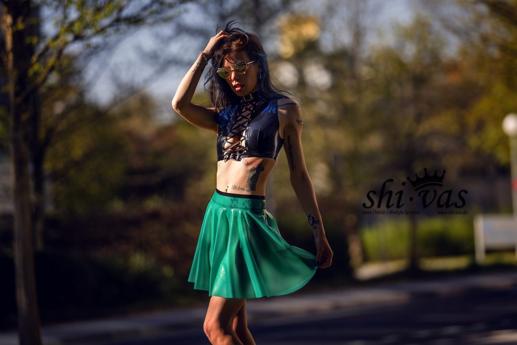 Coco steht im Sonnenlicht auf einer ruhigen Straße, trägt ein schwarzes geschnürtes Latex-Top und einen semi-transparenten grünen Latexrock ohne Slip. Sie spielt mit dem Rocksaum, Latex schimmert im Licht, High Heels, entspannte Pose, selbstbewusste Ausstrahlung.