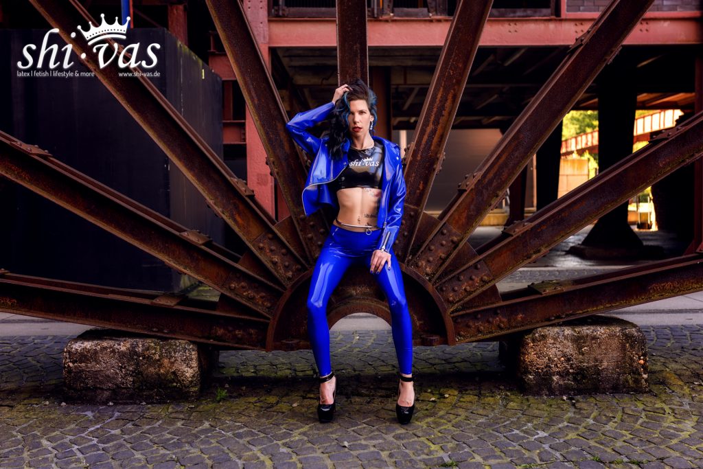 Coco posiert selbstbewusst in glänzendem blauem Latex-Outfit vor massiven Stahlstrukturen, eine Hand im Haar, High Heels auf Kopfsteinpflaster, direkter Blick und starke Körperspannung.