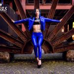 Coco steht selbstbewusst in glänzendem blauem Latex-Outfit zwischen massiven Stahlstrukturen, Arme ausgebreitet, High Heels auf Kopfsteinpflaster, starke Präsenz vor industrieller Kulisse.