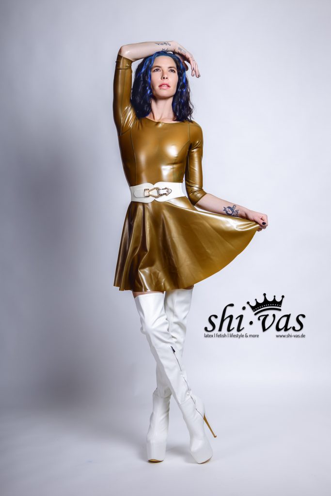 Coco posiert in ihrem goldenen Latexkleid, ein Arm ist über den Kopf angewinkelt, die andere Hand hält den Rock. Sie trägt weiße Overknee-Stiefel mit extremem Absatz. Ihr blau-schwarzes Haar ist lockig gestylt.
