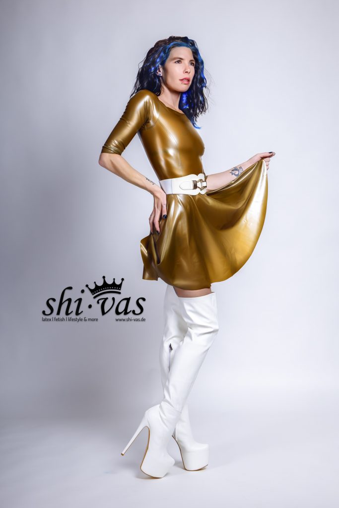 Seitenansicht von Coco im goldenen Latexkleid. Sie hat eine Hand in die Taille gestützt und blickt über die Schulter. Die weißen Overknee-Stiefel sind deutlich sichtbar.