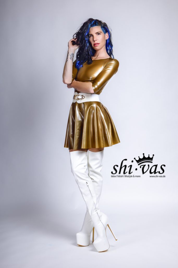 Coco steht in einer Drei-Viertel-Ansicht, die Arme vor der Brust verschränkt, eine Hand spielt an ihrem Haar. Der Fokus liegt auf dem glänzenden Latexkleid und der schlanken Taille.