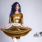 Eine Querformat-Aufnahme von Coco, die frontal zur Kamera steht und mit beiden Händen den Saum ihres goldglänzenden Latex-Skaterkleids weit ausbreitet. Sie trägt einen markanten weißen Taillengürtel, weiße Overknee-Stiefel und ihre Tätowierungen an den Unterarmen sind deutlich sichtbar. Ihr blau-schwarzes Haar fällt lockig über ihre Schulter.