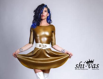 Eine Querformat-Aufnahme von Coco, die frontal zur Kamera steht und mit beiden Händen den Saum ihres goldglänzenden Latex-Skaterkleids weit ausbreitet. Sie trägt einen markanten weißen Taillengürtel, weiße Overknee-Stiefel und ihre Tätowierungen an den Unterarmen sind deutlich sichtbar. Ihr blau-schwarzes Haar fällt lockig über ihre Schulter.