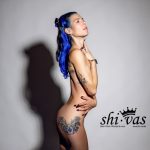 Coco splitternackt im Studio, seitliche Pose mit betonter Silhouette. Sichtbare Tattoos an Rücken und Hüfte, nackte Brüste mit Nippelpiercings, eine Hand am Hals, die andere an der Taille. Neutraler Hintergrund, sinnliche Haltung.