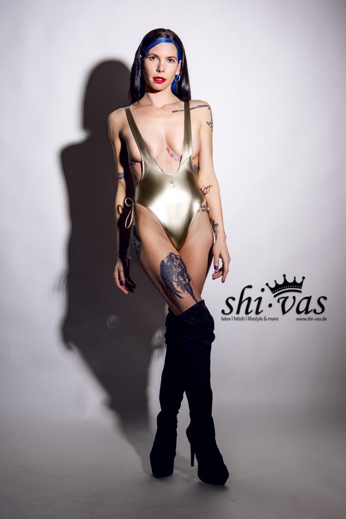 Coco steht frontal, Arme hängen locker an der Seite, leichtes Lächeln, der goldene Anzug spannt sich über ihren Körper.