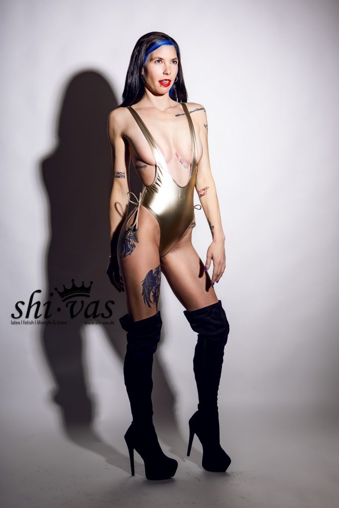 Coco steht in voller Größe, ein Bein leicht angewinkelt, Arme hängen locker herab, der Anzug spannt sich extrem über den Schritt.