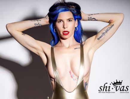 Querformatige Nahaufnahme von Coco, die beide Hände hinter ihren Kopf legt und direkt in die Kamera blickt. Sie trägt einen goldenen Badeanzug, hat blaue Haare und Lippenpiercings.