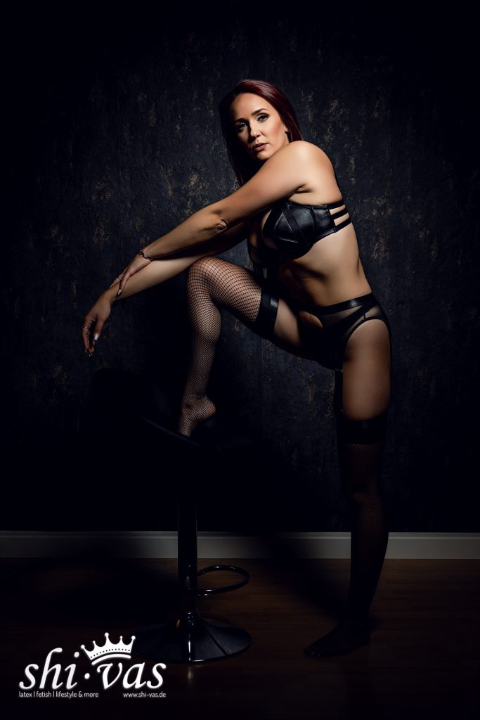 Finnja steht neben dem Hocker, ein Bein darauf abgestützt, den Oberkörper nach vorne gebeugt. Ihr Blick ist fixiert auf die Kamera, die Hand ruht auf dem Knie.