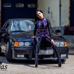 Gastmodel Amy Grey sitzt im schwarzen Latex-Catsuit mit violettem Harness auf der Motorhaube eines dunklen BMW vor einer industriellen Kulisse
