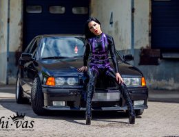 Gastmodel Amy Grey sitzt im schwarzen Latex-Catsuit mit violettem Harness auf der Motorhaube eines dunklen BMW vor einer industriellen Kulisse