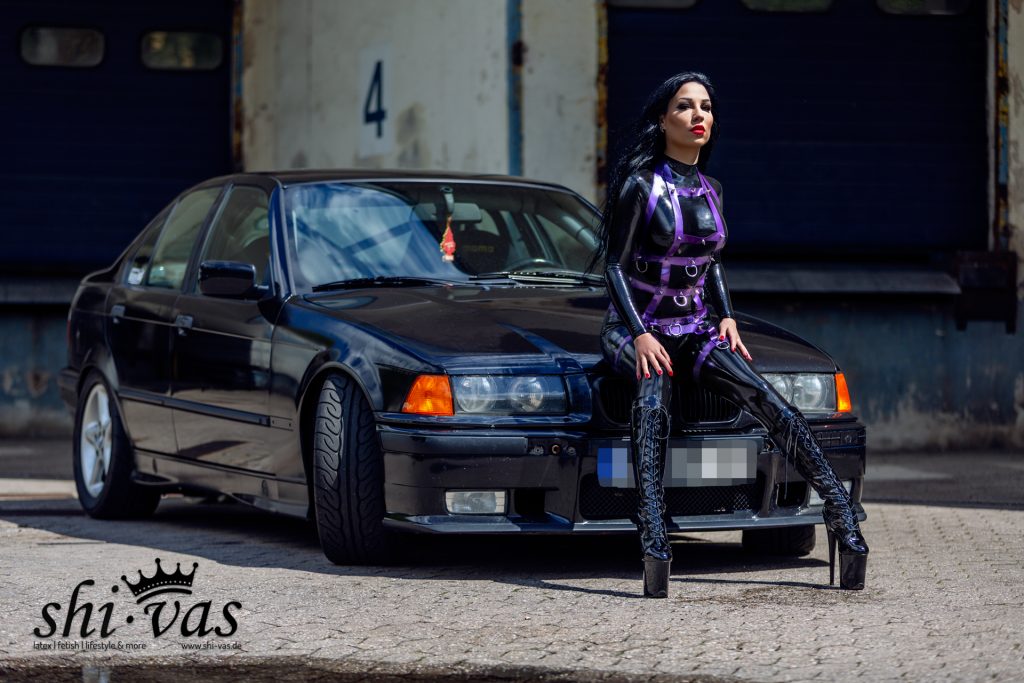 Gastmodel Amy Grey sitzt im schwarzen Latex-Catsuit mit violettem Harness auf der Front eines dunklen BMW in industrieller Umgebung