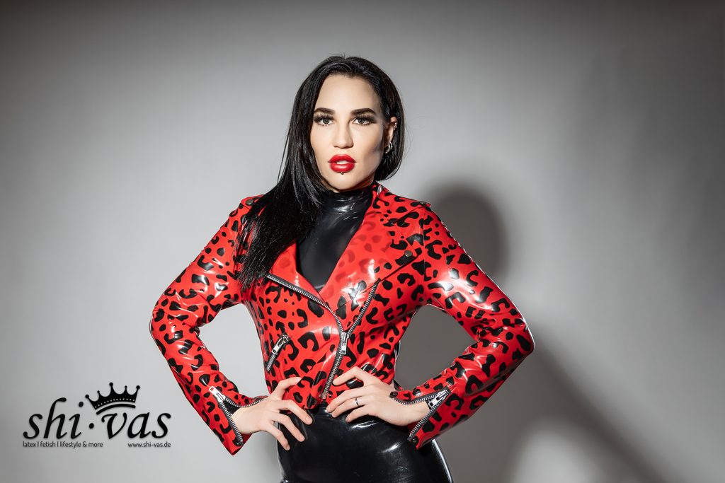 Amy Grey steht frontal vor grauem Hintergrund, die Hände in die Hüften gestützt. Sie trägt eine rote Latex-Bikerjacke mit schwarzem Leopardenmuster über einem schwarzen Latex-Body.