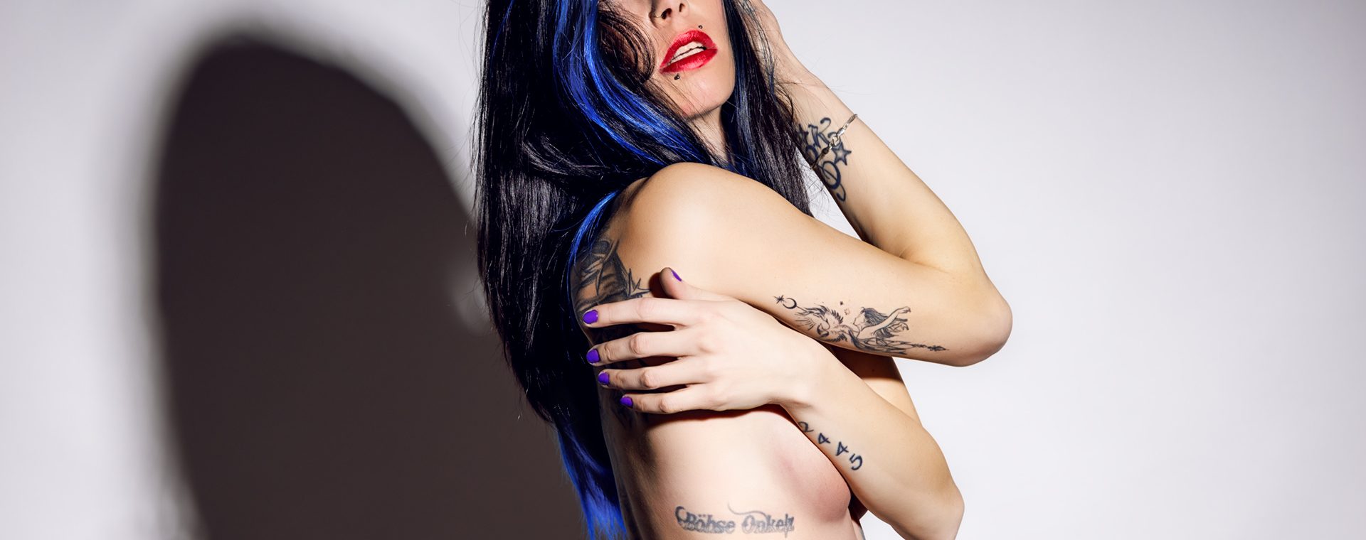 Seitliche Studioaufnahme von Coco mit geöffnetem goldenen Badeanzug, Hand im Haar, zweite Hand am Arm, direkte Blickführung, starker Schlagschatten.