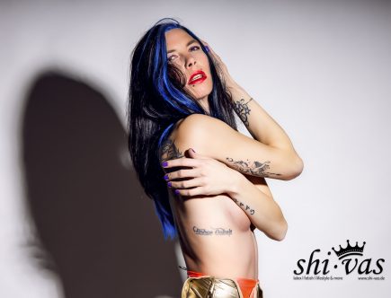 Seitliche Studioaufnahme von Coco mit geöffnetem goldenen Badeanzug, Hand im Haar, zweite Hand am Arm, direkte Blickführung, starker Schlagschatten.