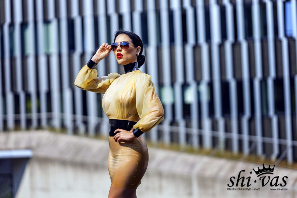 Amy Grey rückt sich die Sonnenbrille zurecht. Das gelbe Latex glänzt stark, der schwarze Kragen betont ihren Hals.