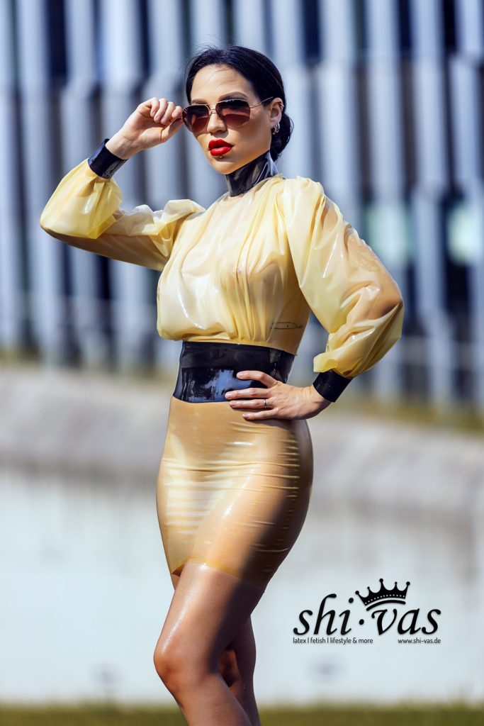 Nahaufnahme von Amy Grey im Profil. Das gelbe, semitransparente Latex der Bluse schimmert im Sonnenlicht und lässt die Silhouette des Oberkörpers erahnen.