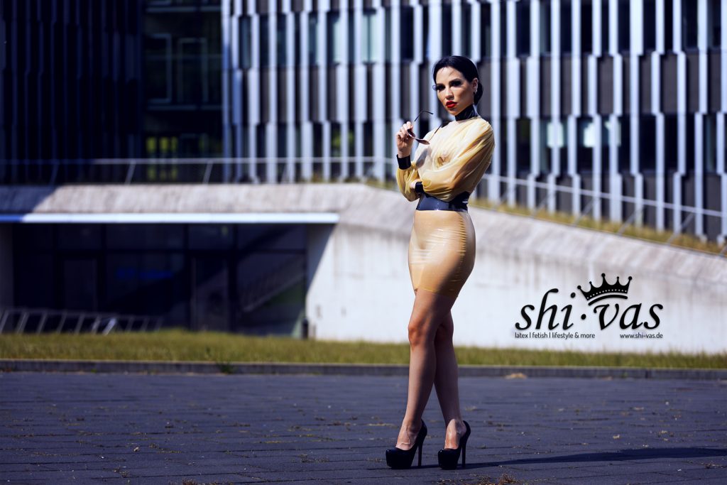 Amy Grey steht in voller Länge auf dem Platz, die Beine leicht überschlagen, die Sonnenbrille lässig in der Hand.