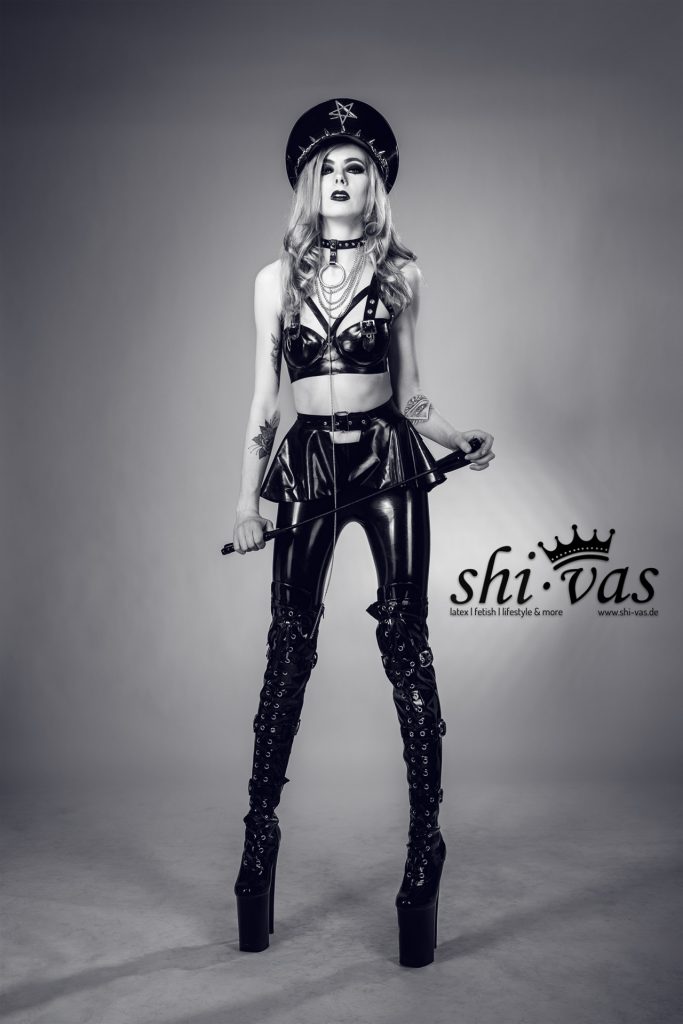 Schwarz-Weiß-Aufnahme von Lena Rawk in einem knappen Latex-Outfit mit Rockschößchen, hoher Offiziersmütze, Halsband und Gerte in den Händen.