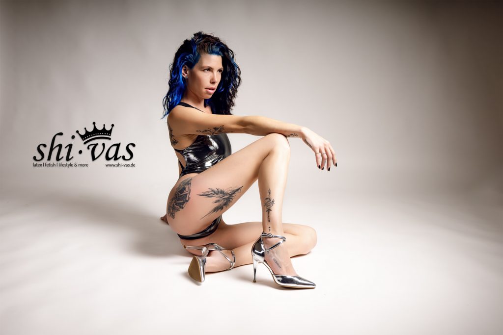 Coco sitzt seitlich auf dem Boden im silbernen, stark reflektierenden LEOHEX-Swimsuit und silbernen High Heels, ein Bein angewinkelt, neutraler Studiohintergrund