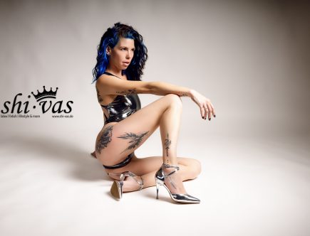 Coco sitzt seitlich auf dem Boden im silbernen, stark reflektierenden LEOHEX-Swimsuit und silbernen High Heels, ein Bein angewinkelt, neutraler Studiohintergrund