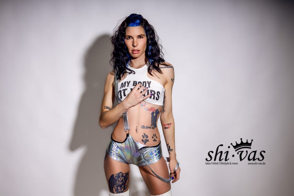 Querformat. Coco frontal, Beine auseinander. Rechte Hand auf dem Top/Brust, linke Hand hält locker einen silbernen Hosenträger. Direkter Blick in die Kamera.