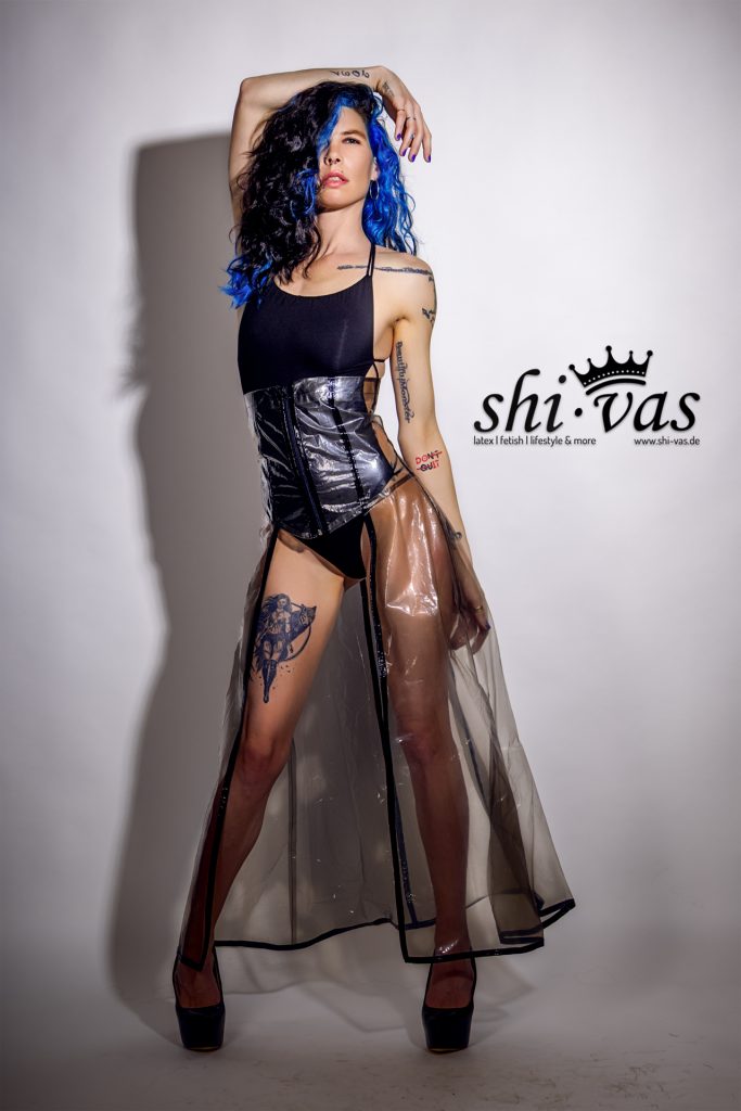 Coco steht in einer weitbeinigen Pose, den rechten Arm über den Kopf gehoben. Der transparente Rock fällt weit, ihre Tattoos an den Armen und Beinen sind in der vollen Länge sichtbar.