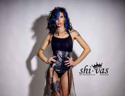 Halbkörperaufnahme von vorne. Coco hat beide Hände fest in die Taille gestützt, was die Silhouette des schwarzen Bodys betont. Ihr Blick ist herausfordernd, die blau gefärbten Haare fallen wild über das Gesicht.