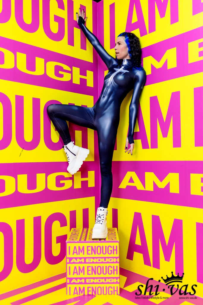 Coco posiert in einem glänzenden, dunkelblauen Ganzkörperanzug vor einer gelb-pinken Wand mit dem Aufdruck „I AM ENOUGH“. Ein Bein hat sie auf eine gleichfarbige Box gestellt, ein Arm ist nach oben gestreckt. Sie trägt weiße Plateau-Boots.