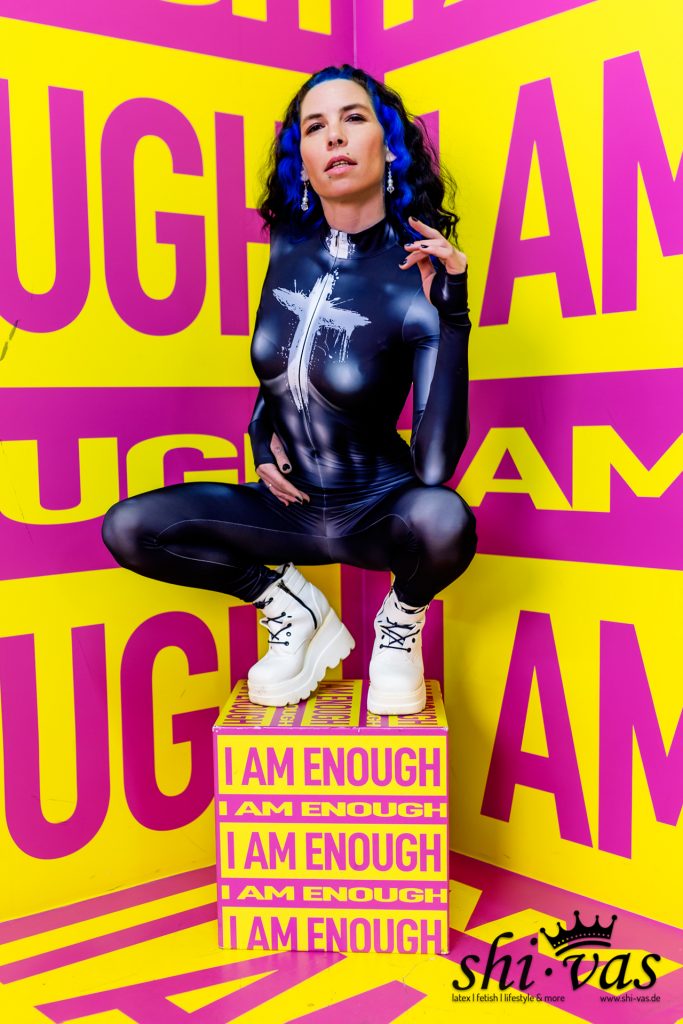 Nahaufnahme von Coco, die auf einer Box hockt. Sie blickt mit leicht geöffnetem Mund direkt in die Kamera. Ihre blauen Haare und die langen Ohrringe sind deutlich sichtbar, ebenso wie der Glanz des blauen Stoffes auf ihrer Haut.