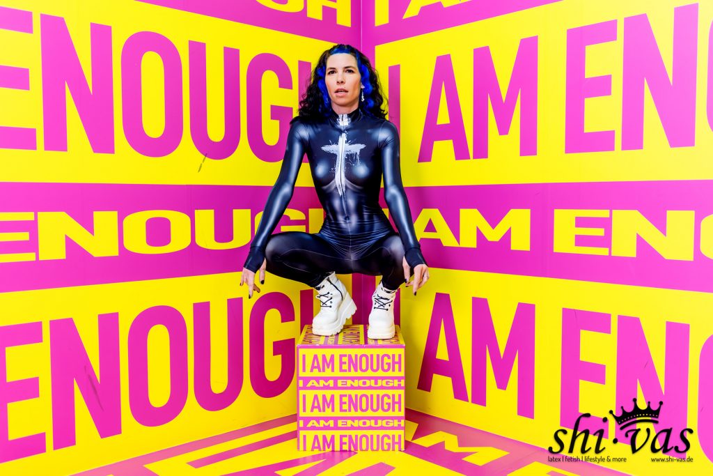 Frontalansicht von Coco in tiefer Hocke auf einer Box. Der dunkelblaue Anzug betont ihren Körper, auf der Brust ist ein weißes, kreuzähnliches Muster zu sehen. Der Hintergrund besteht aus knalligen gelben und pinken Textflächen.