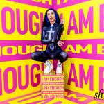 Coco hockt auf einer gelben Box, die mit „I AM ENOUGH“ beschriftet ist. Sie trägt einen dunkelblauen Catsuit und weiße Plateau-Schuhe. Ihre Arme sind vor dem Körper verschränkt, die Finger gespreizt.